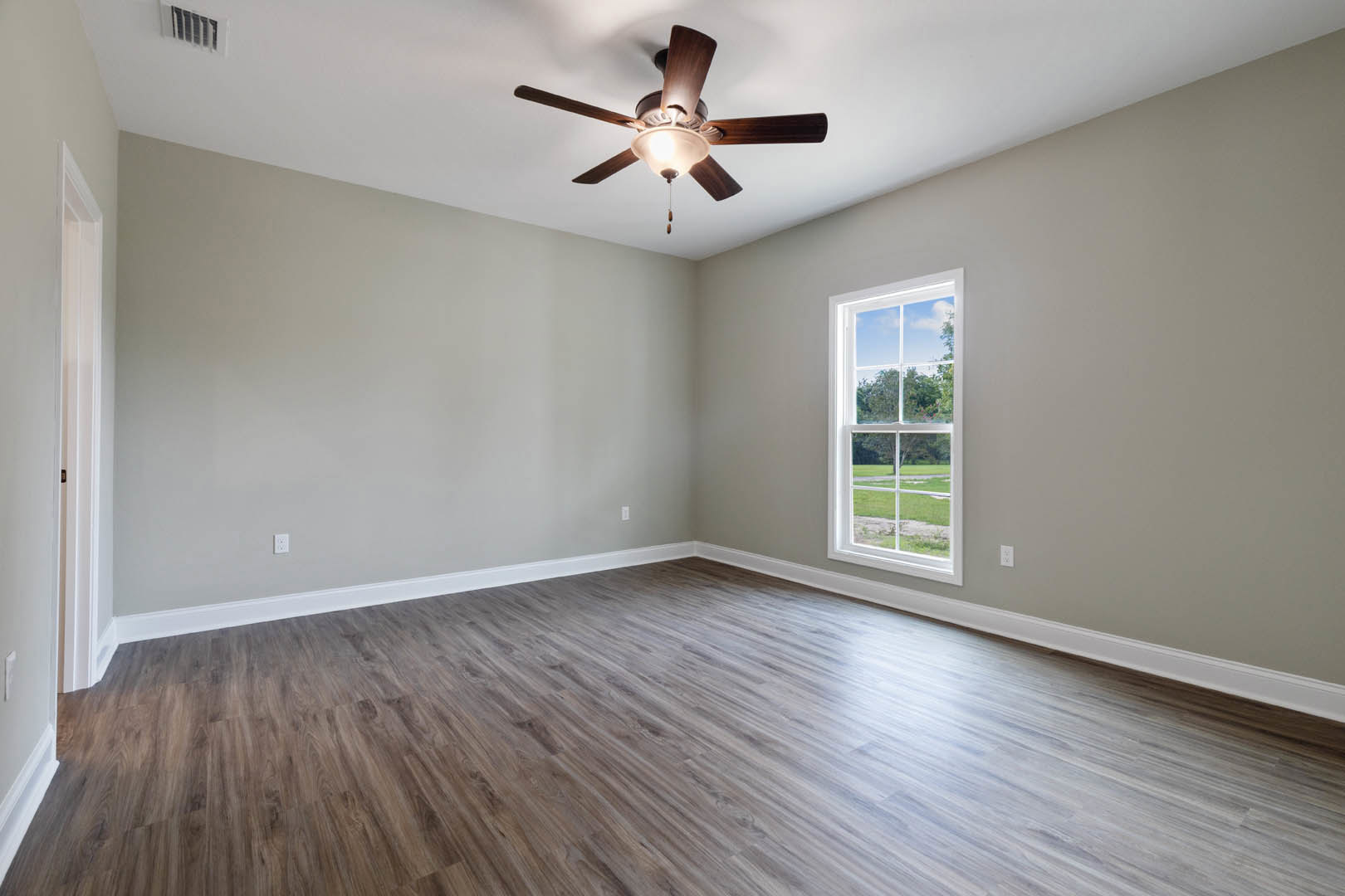 Ceiling fan with integrated light fixture mounted on white plaster ceiling, large window overlooking green lawn, wood flooring, neutral painted walls