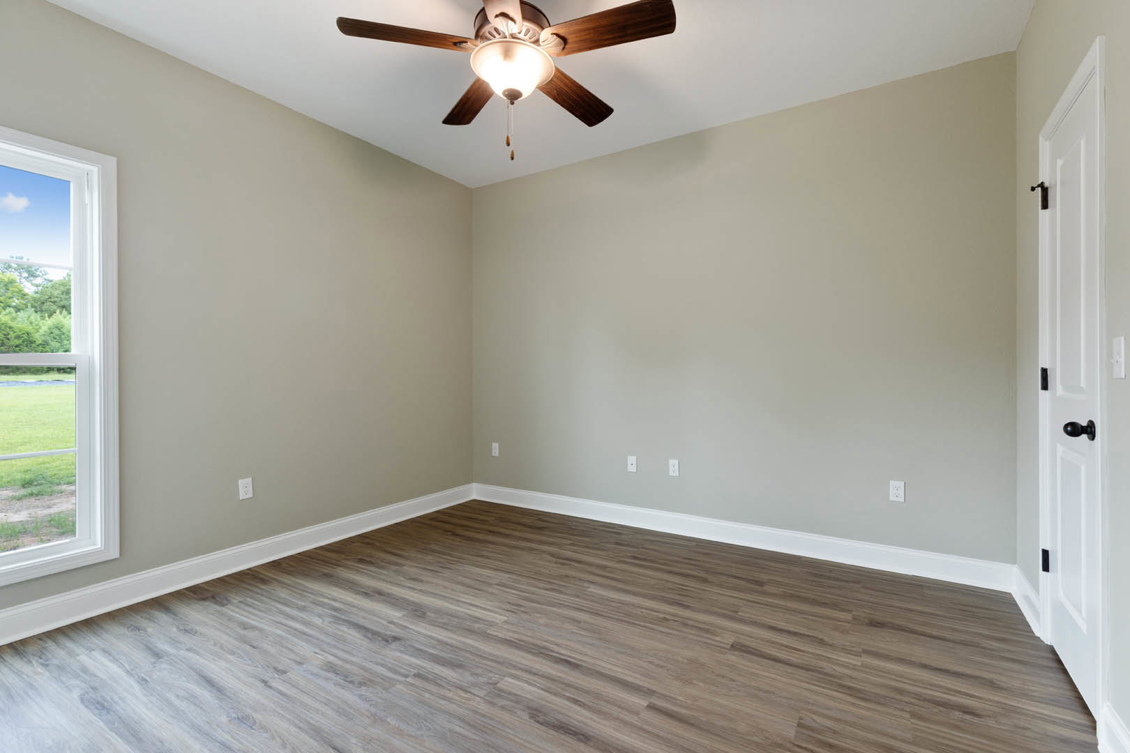Ceiling fan with light fixture centered above wood flooring and white baseboards, window overlooking grassy field, neutral walls and molding.
