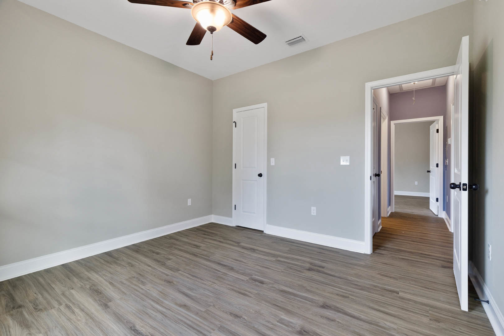 Ceiling fan with light fixture mounted on white plaster ceiling, wood laminate flooring, white door with black handle and knobs, neutral walls.