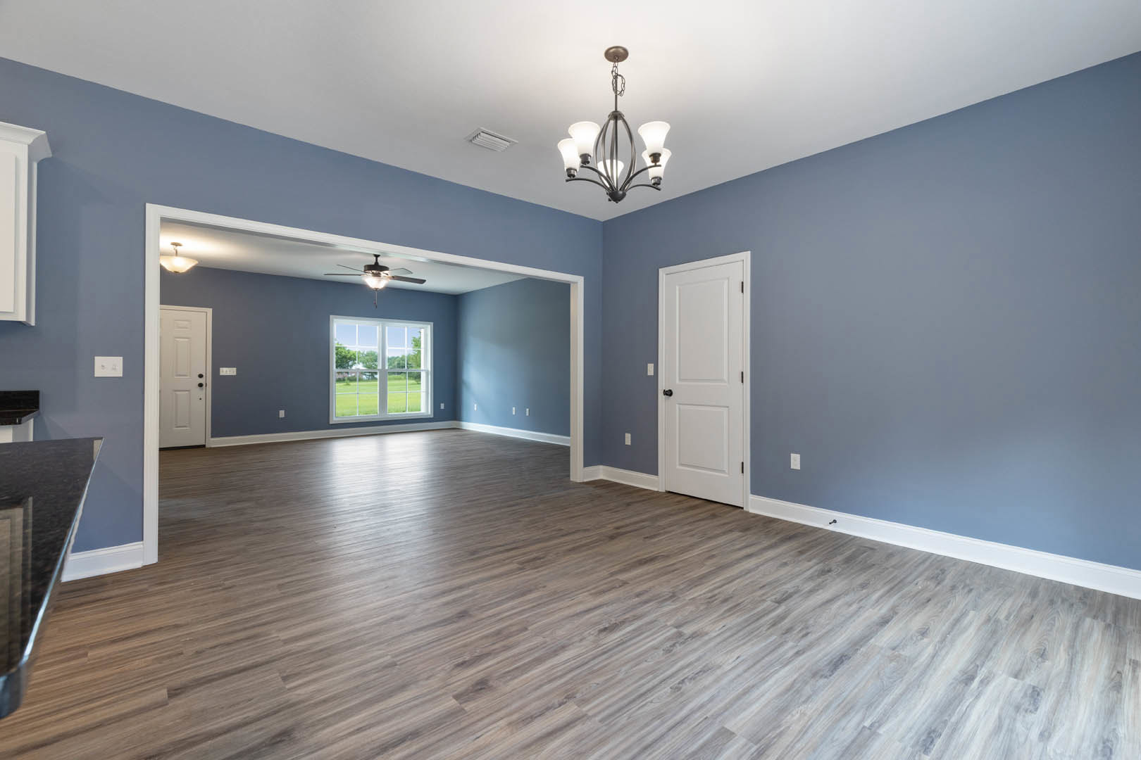 Wood flooring in a bright room with a ceiling fan, white walls, black hardware on a white door, large window overlooking a green lawn, and a close-up of a chandelier.