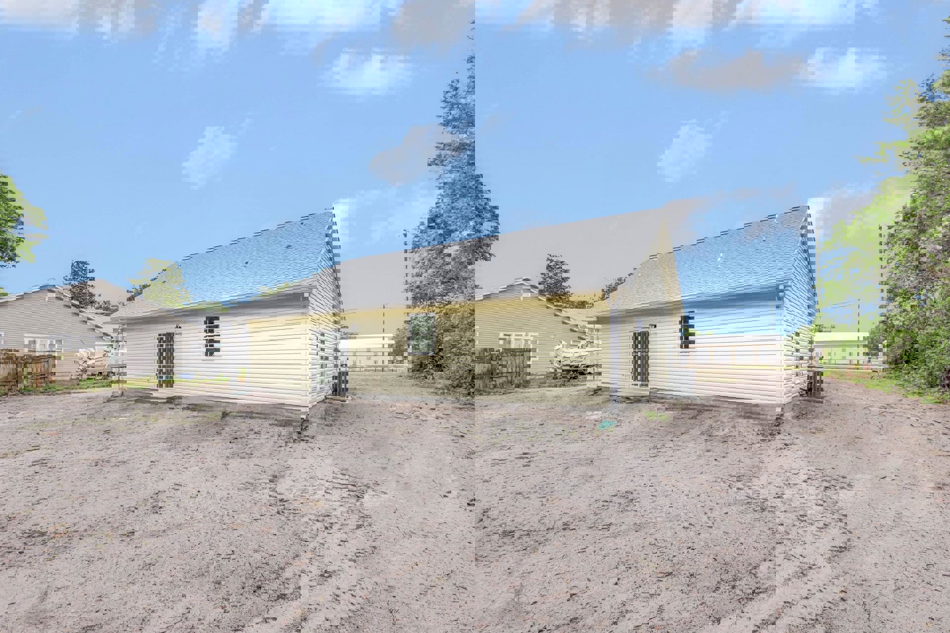 Yellow cottage with open front door, white-trimmed windows, multi-pane glass, dirt yard featuring a green object, wooden fence lined with plants, cloudy sky overhead.