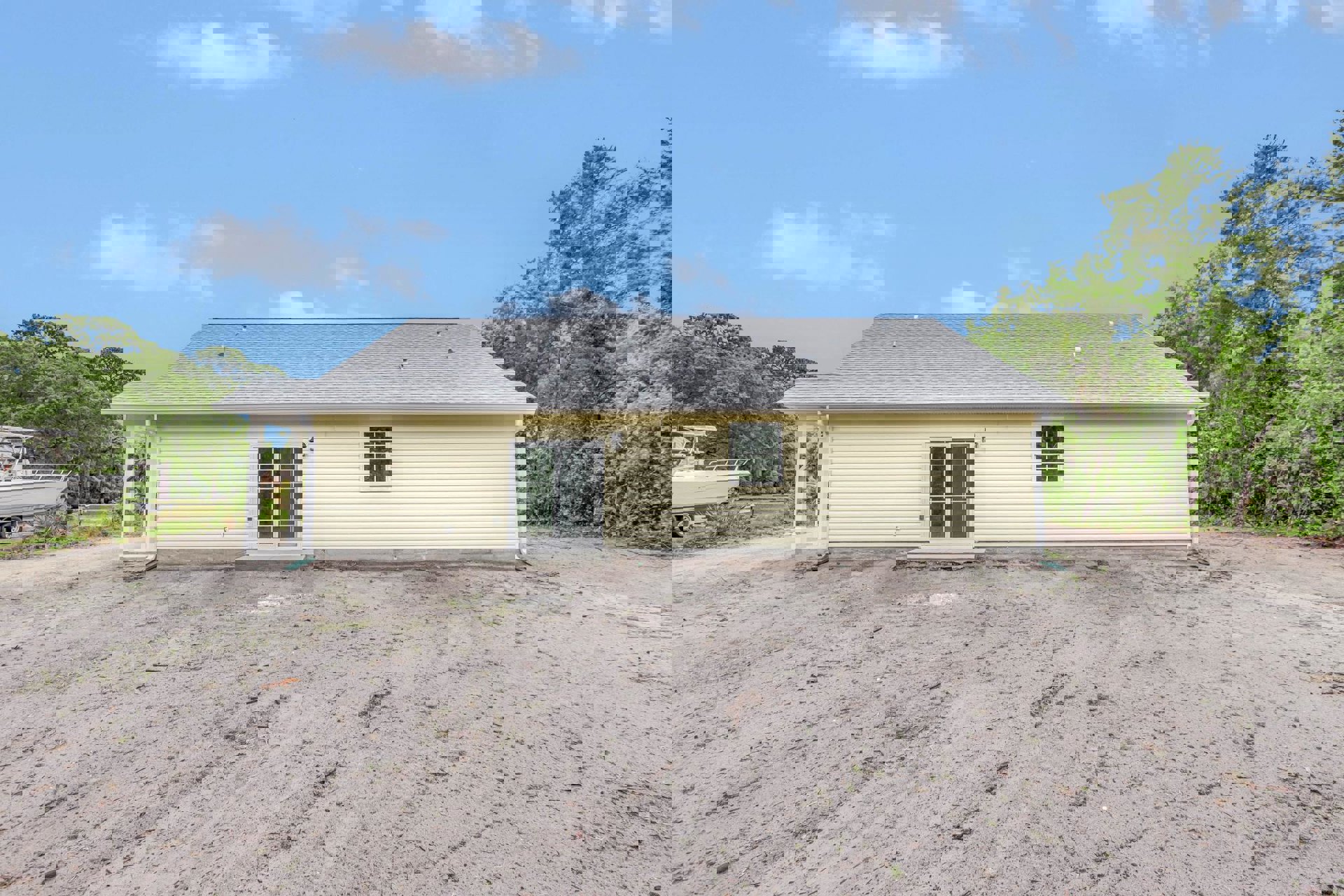 Yellow cottage-style home with white roof, white-framed windows, white sliding glass door, boat on trailer parked beside dirt yard, surrounded by trees under partly cloudy sky
