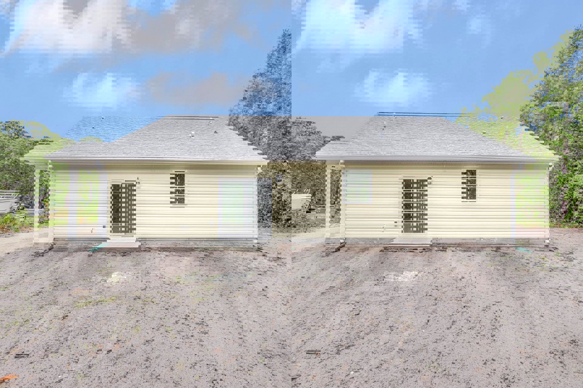 Single-story home with white-framed windows and sliding glass door, dirt driveway and yard, blue sky with scattered clouds overhead