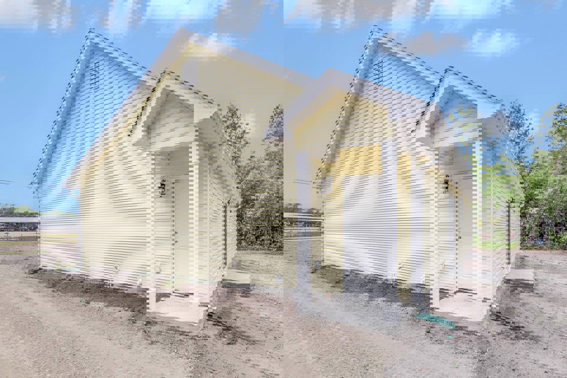 Yellow house with white door, white columns, and white wall vent, set along a dirt road under a blue sky with scattered clouds.