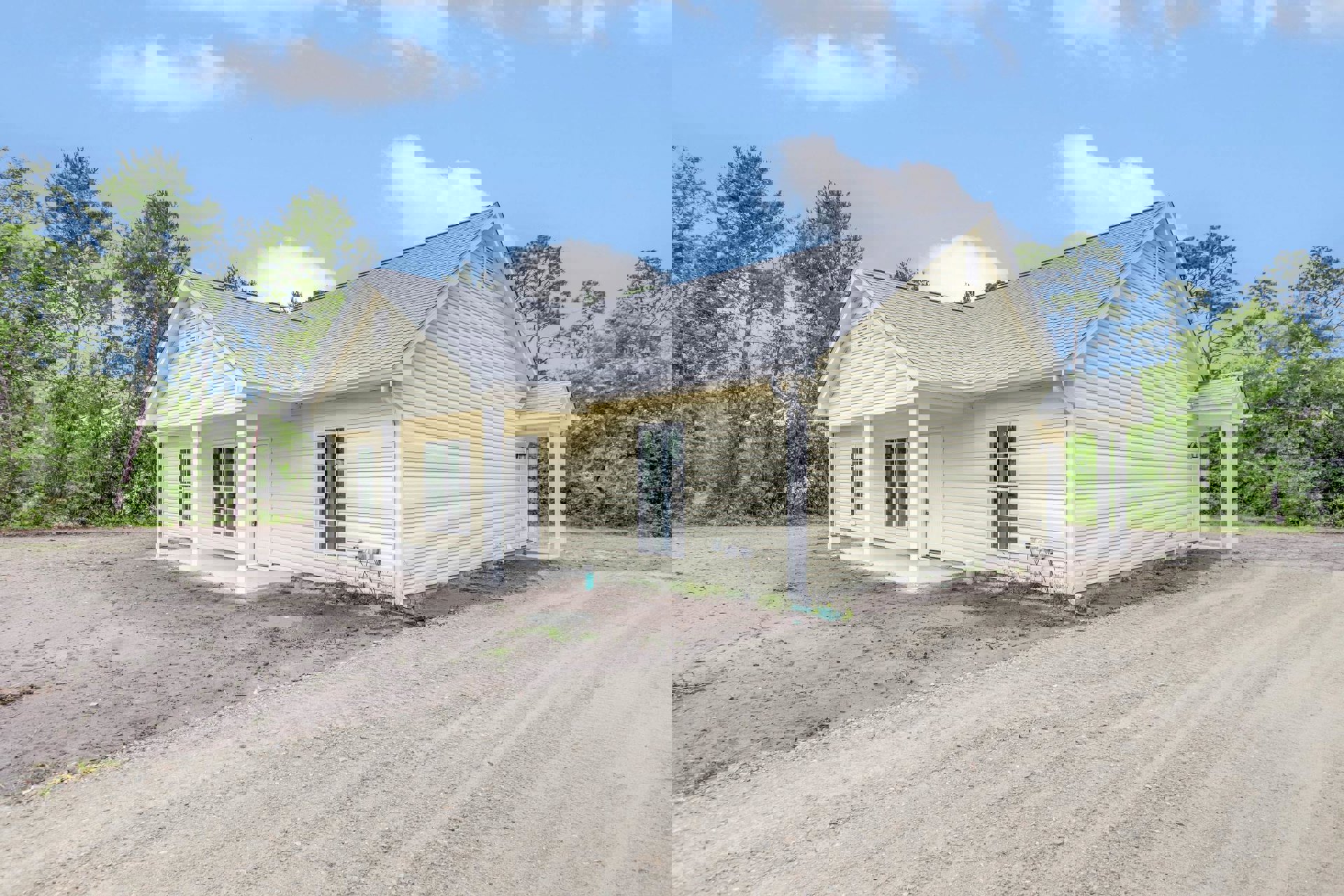 Yellow cottage-style home with multi-pane white windows, white front door, dirt road running alongside, surrounded by mature trees under a partly cloudy blue sky