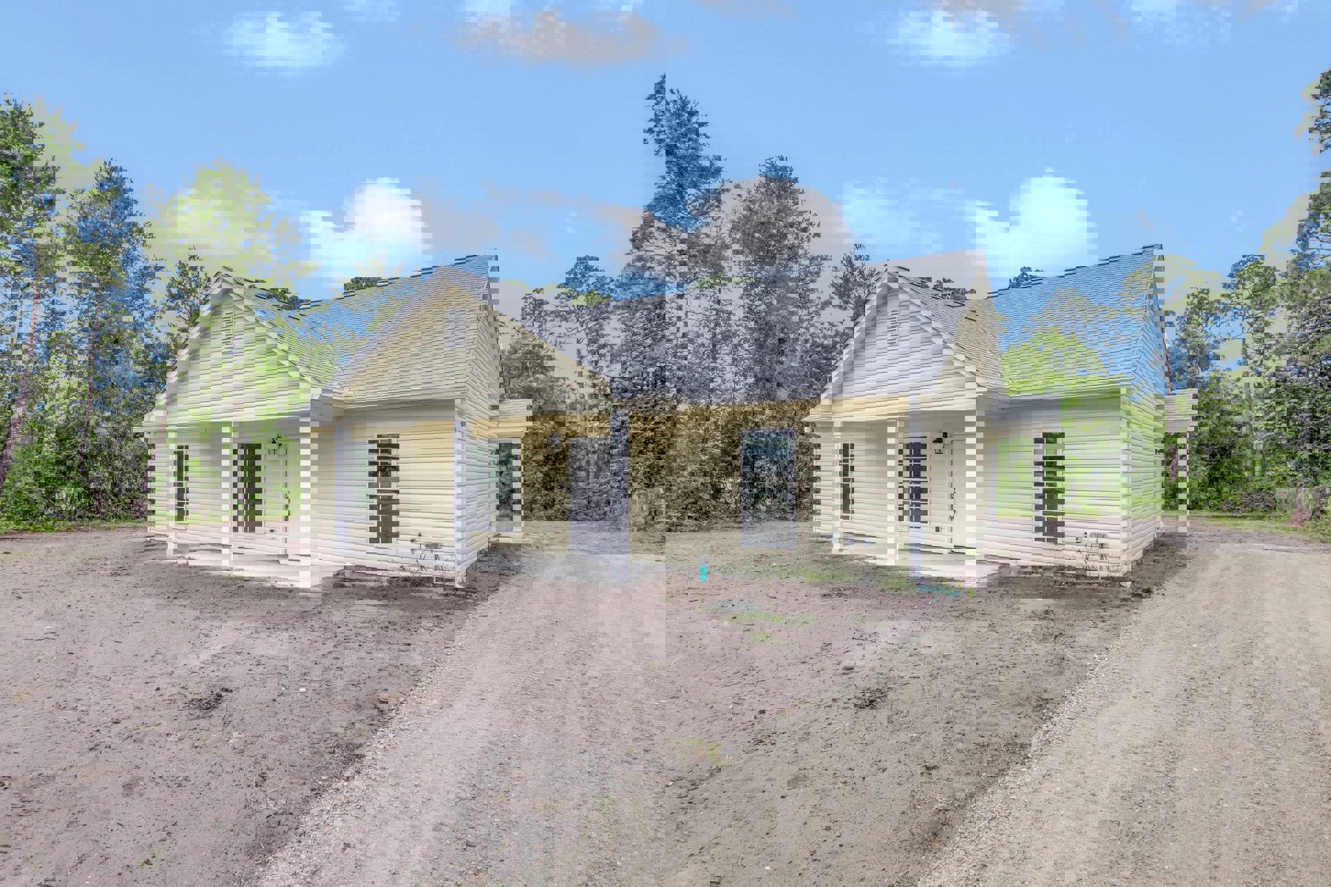 Two-story house with white door, glass panes, white-framed window, light fixture, dirt road and patch in front, surrounded by trees and plants under cloudy sky