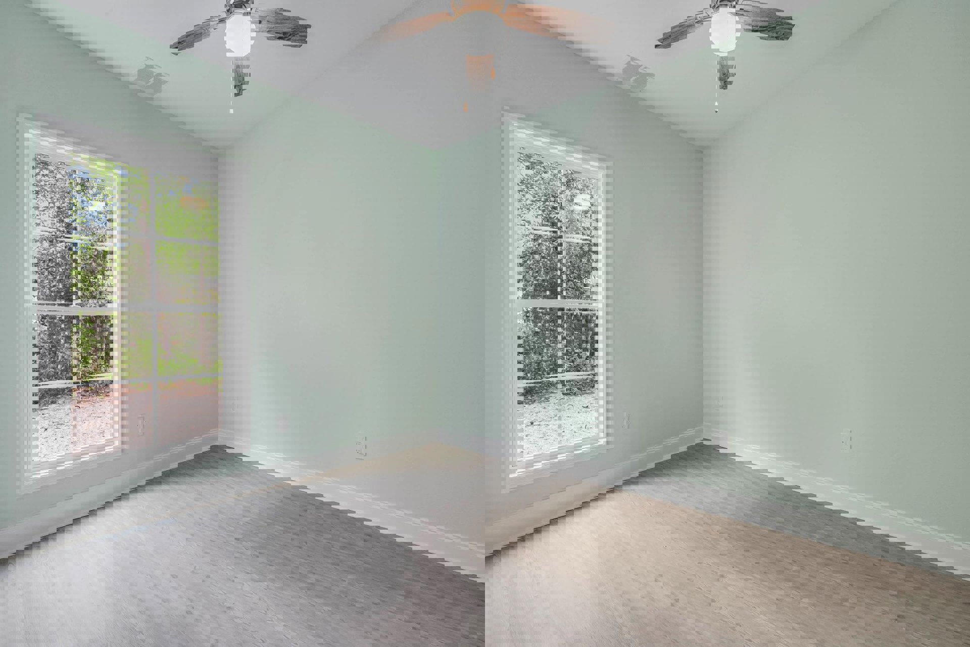 Wood floor room with white walls, ceiling fan with light fixture, large window showing trees outside, ceiling vent, gold-framed circular decor.