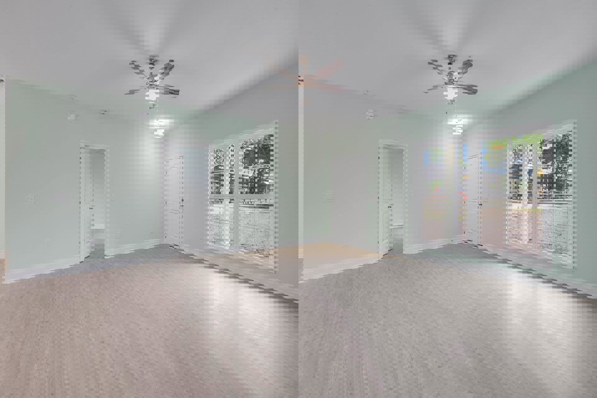 Ceiling fan with light fixture, wood laminate flooring, white plaster walls, white door with silver handle, window overlooking dirt road