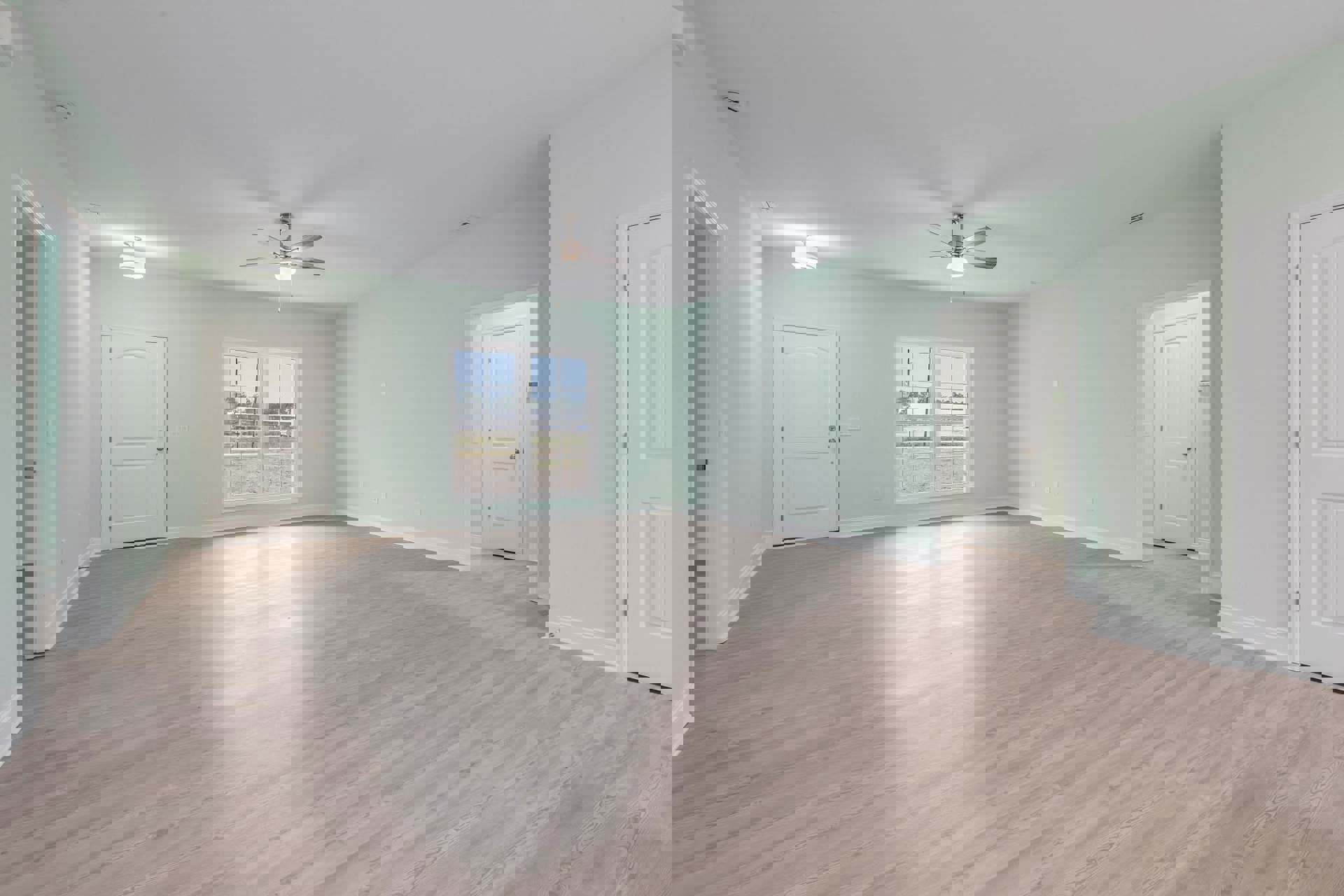 Wood flooring in a bright room featuring a ceiling fan with light, white door with silver handle, window overlooking a grassy field, and neutral plaster walls.