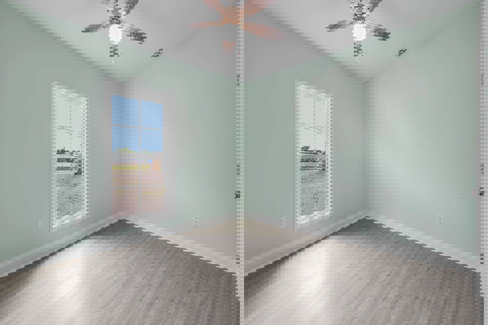 Ceiling fan with light fixture above wood flooring, large window showing dirt road and blue sky, white walls with molding, person holding white plate near window