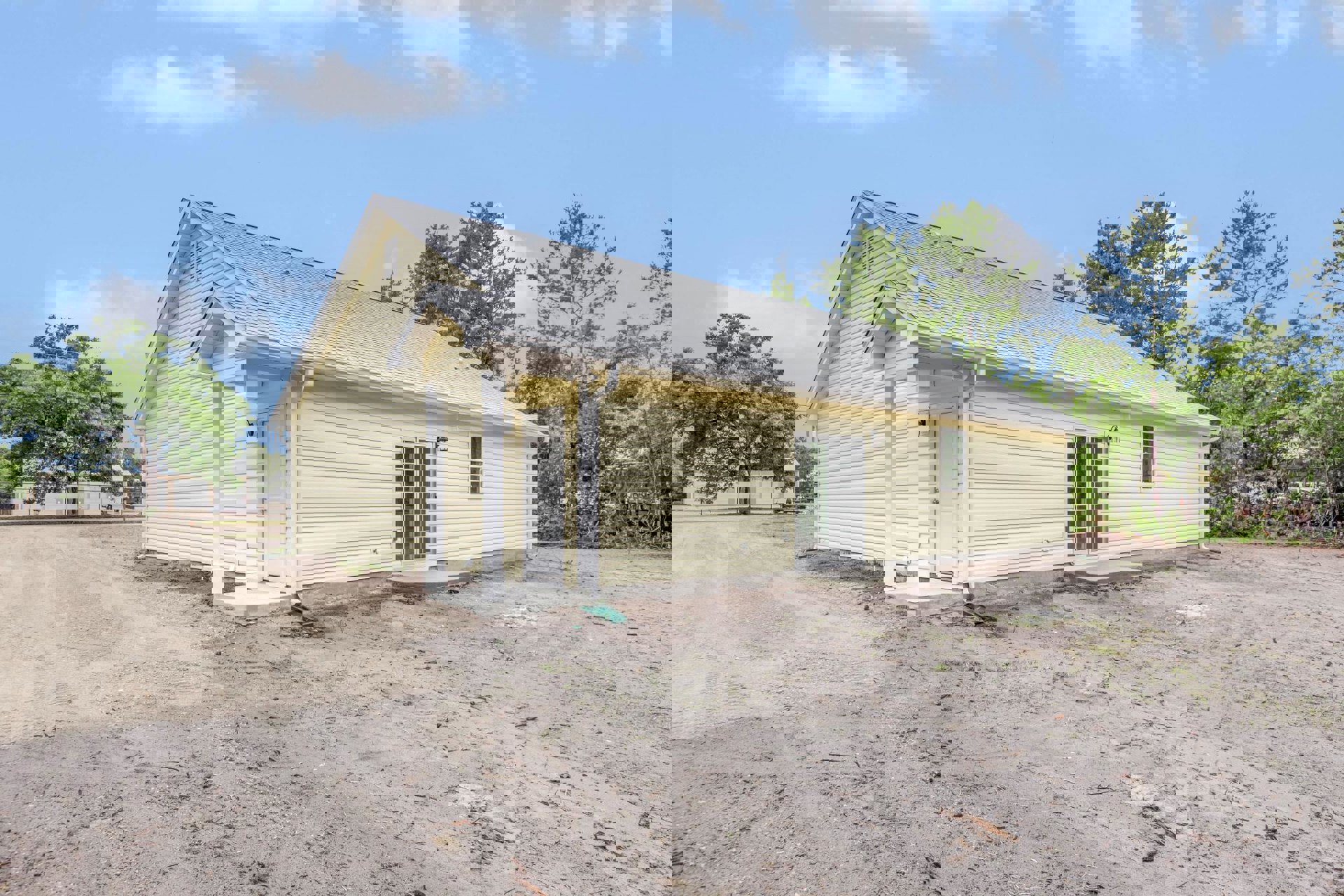 Yellow siding house with white door and glass panes, surrounded by green leafy trees, dirt road and bare ground in front, cloudy sky overhead