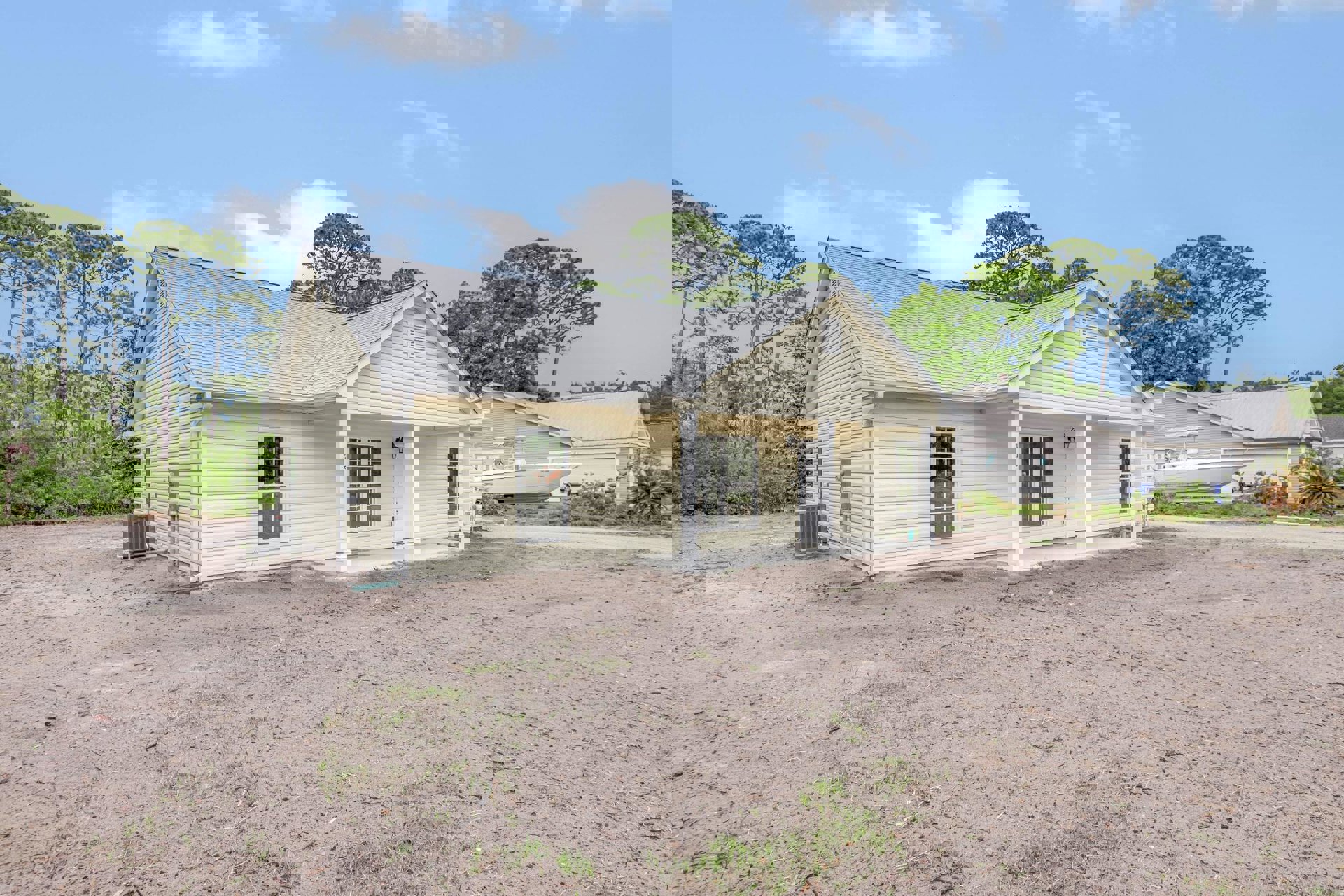 Single-story house with white roof, white-framed windows, and white door with glass panes, set behind a dirt yard with a grey metal box featuring a red handle.