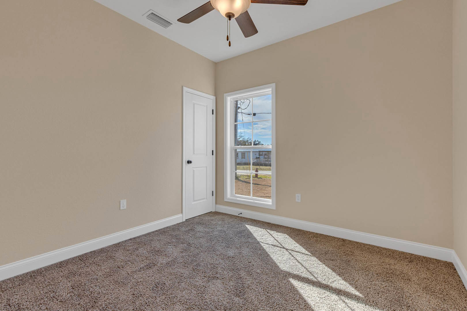Carpeted room with white walls, ceiling fan with light fixture, large window showing neighboring house, white door with black hardware, crown molding along ceiling