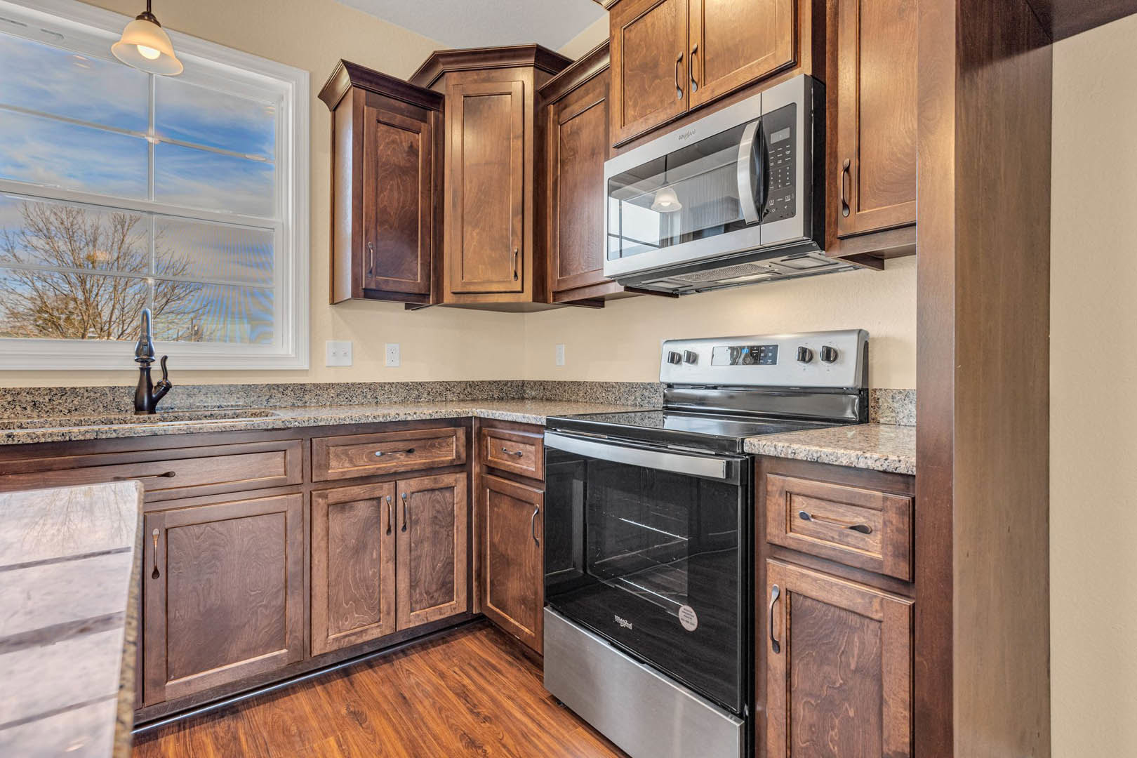 Kitchen with natural wood cabinets, stainless steel stove and microwave, marble countertop, ceiling light fixture, sunlight streaming through window, close-up of wood grain on
