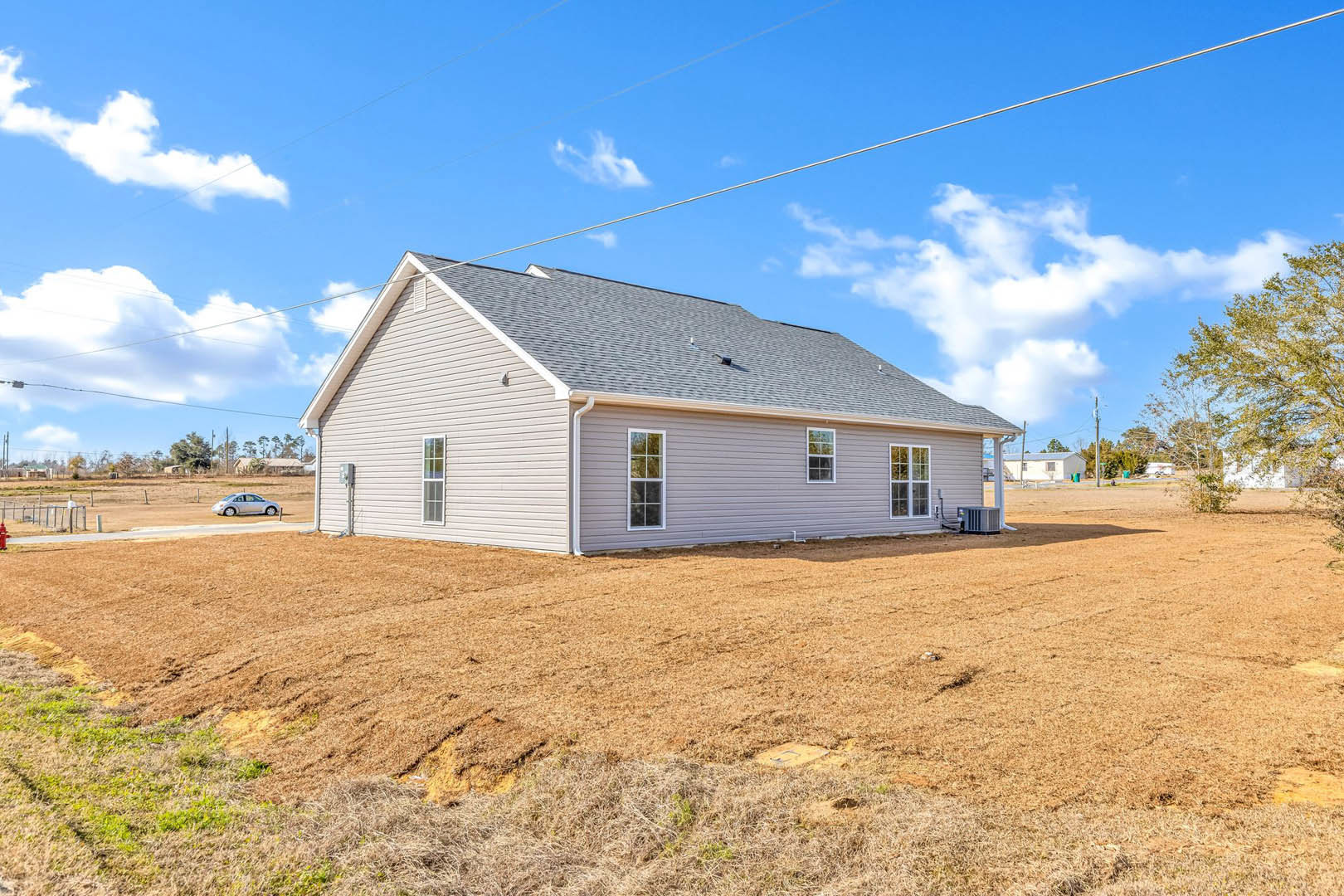 Two-story house with white-framed windows, patch of grass beside a large dirt field, power lines overhead, and trees in the background under a cloudy sky