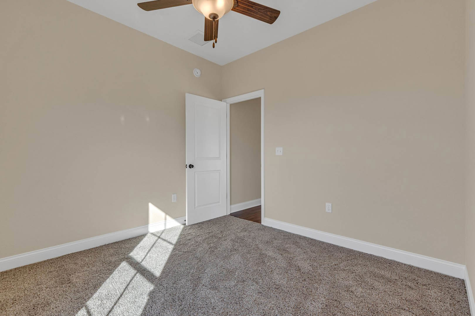 Neutral-toned carpeted room with white walls, ceiling fan featuring a light fixture, white door with black knob, and white door frame molding.