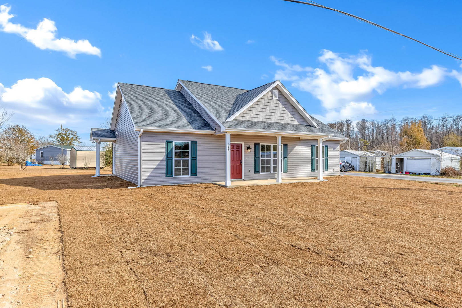 Two-story home with blue shuttered windows, red front door, gray roof, and expansive brown lawn bordered by mature trees under a partly cloudy sky