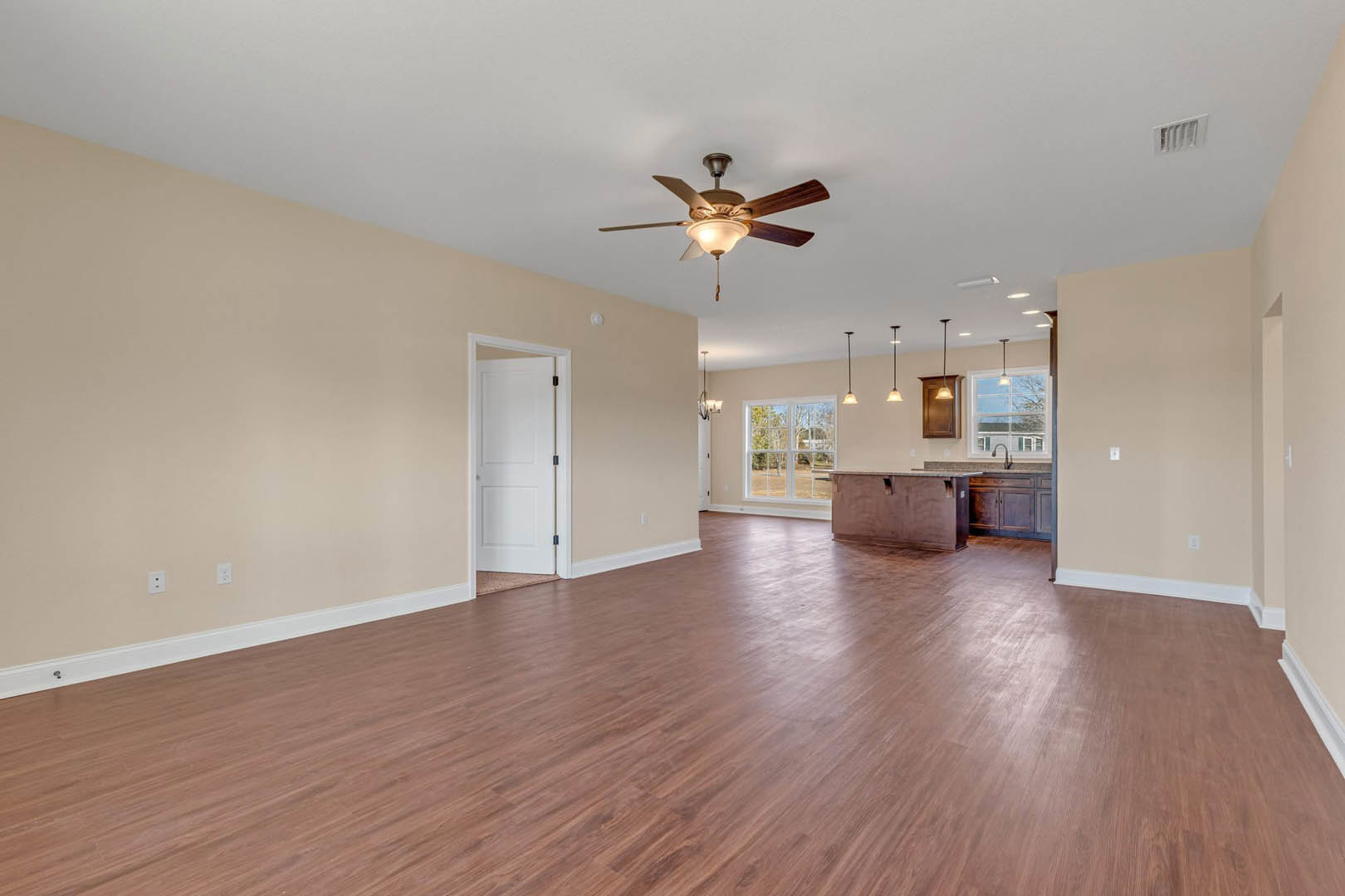 Spacious room featuring hardwood floors, white walls, ceiling fan with light fixture, white door with black handle, wall vent, and a desk in the corner.