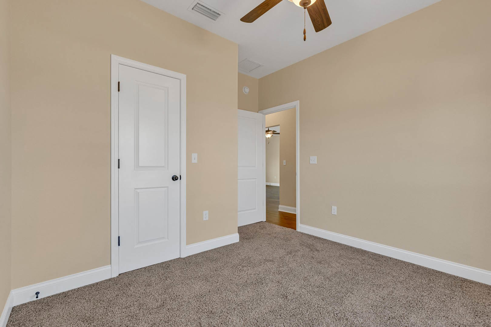 Beige-walled room with plush carpet, white doors featuring black knobs and brown trim, ceiling fan with integrated light fixture, visible light switch on wall