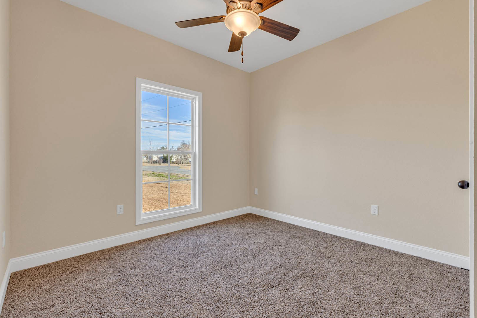Carpeted room with white walls, ceiling fan with light fixture, large window overlooking suburban street and houses, crown molding along ceiling edges