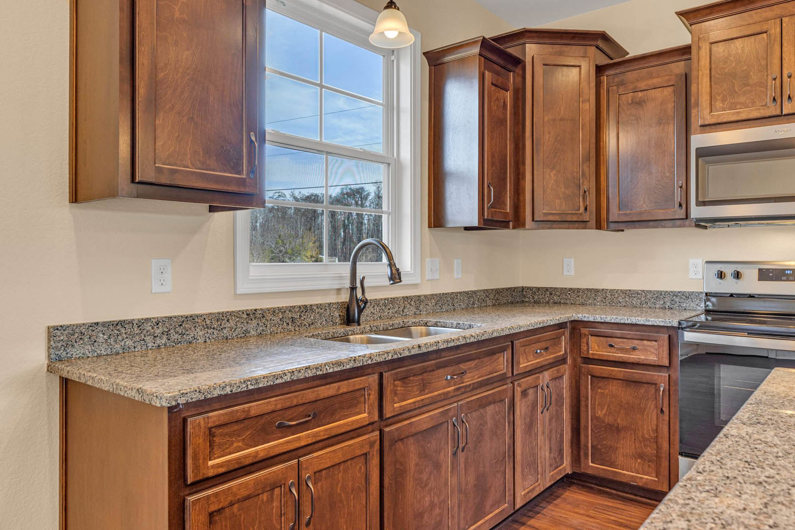 Kitchen featuring wooden cabinets, granite countertops, stainless steel sink with chrome faucet, open microwave, electrical outlet, and window overlooking trees and sky.