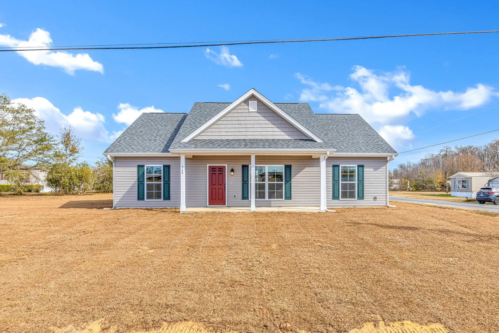 Red front door with white trim on a cottage-style home, blue shuttered windows, brown grass lawn, and cloudy sky in the background.