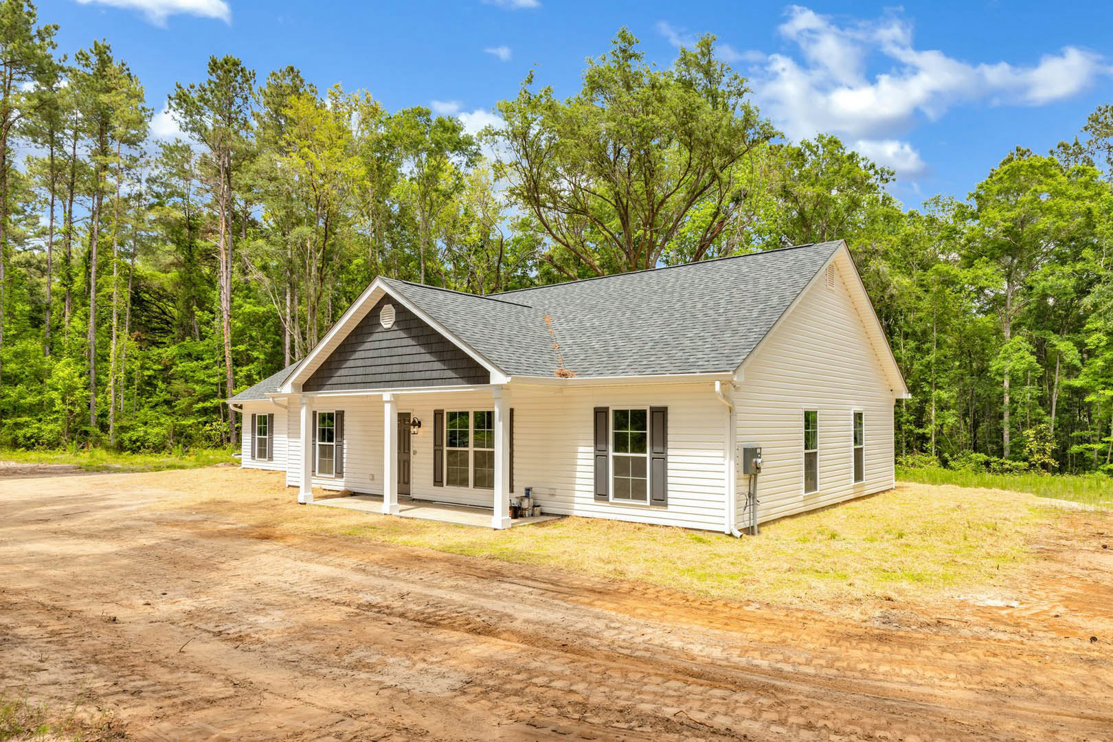 Two-story house with gray roof and covered porch, white-framed windows, dirt road running alongside, surrounded by mature trees and open sky