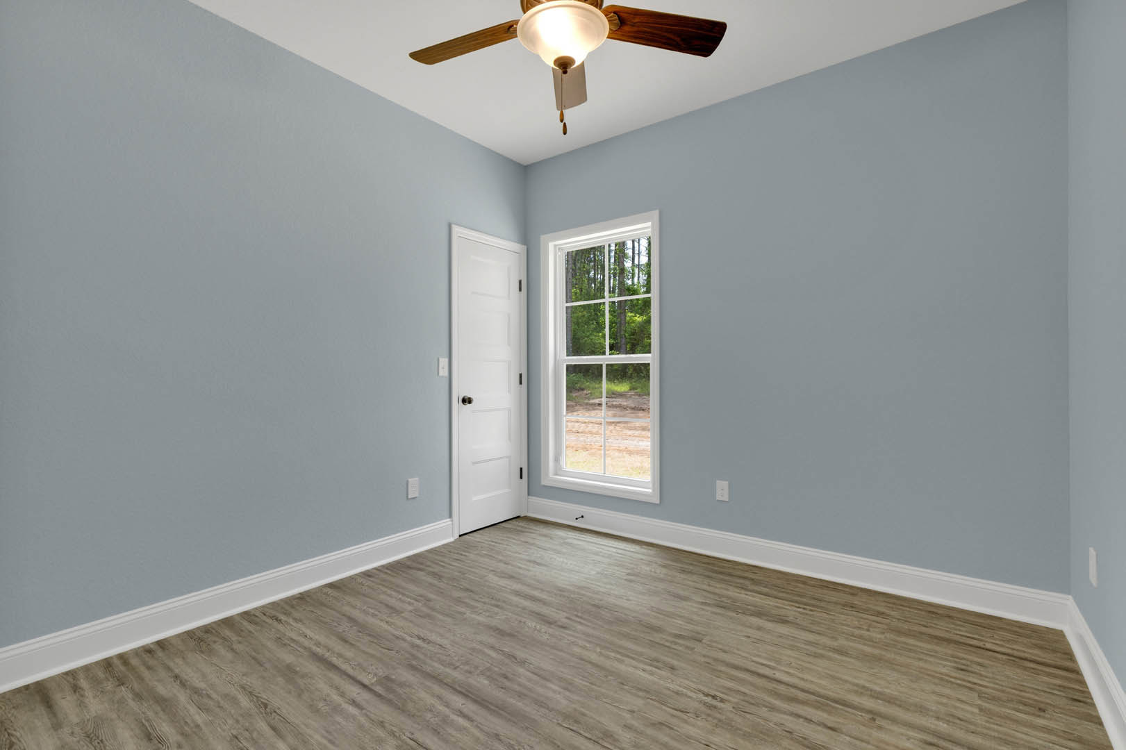 Ceiling fan with light fixture mounted on white plaster ceiling, large window overlooking forest, wood laminate flooring with white baseboard trim, white paneled door with silver