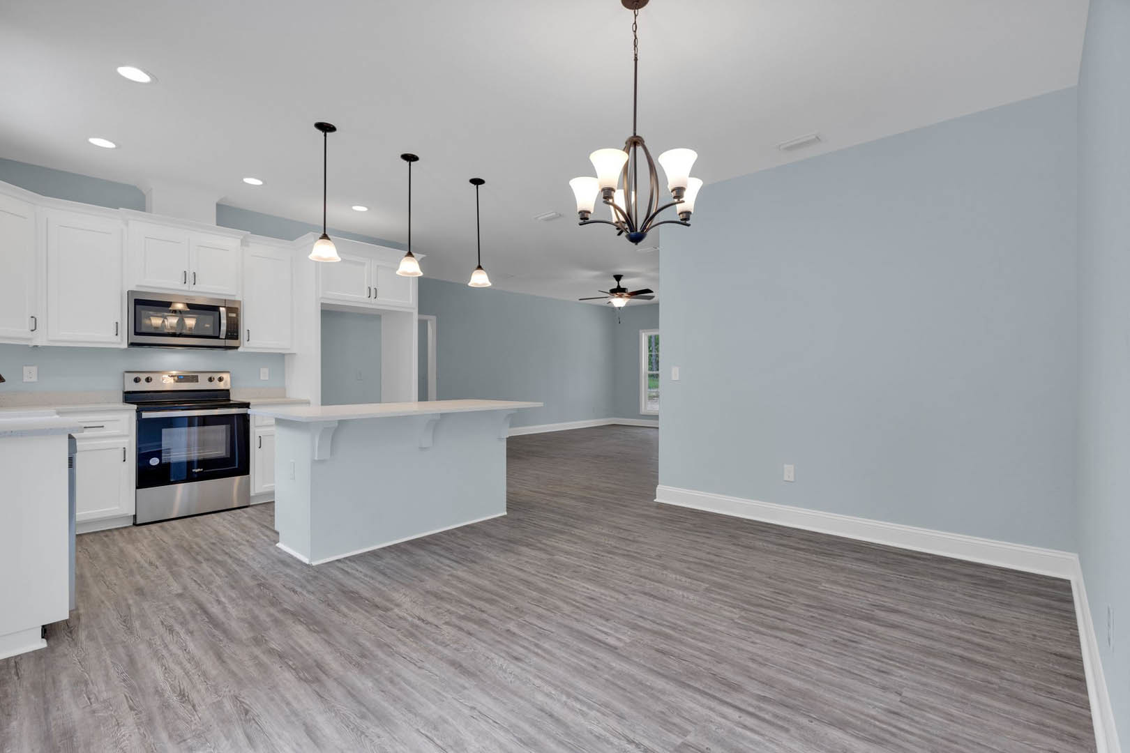 Open kitchen and dining area with wood flooring, white countertops, cabinetry, stainless steel stove and oven, modern chandelier, and blurred lamp in the background.