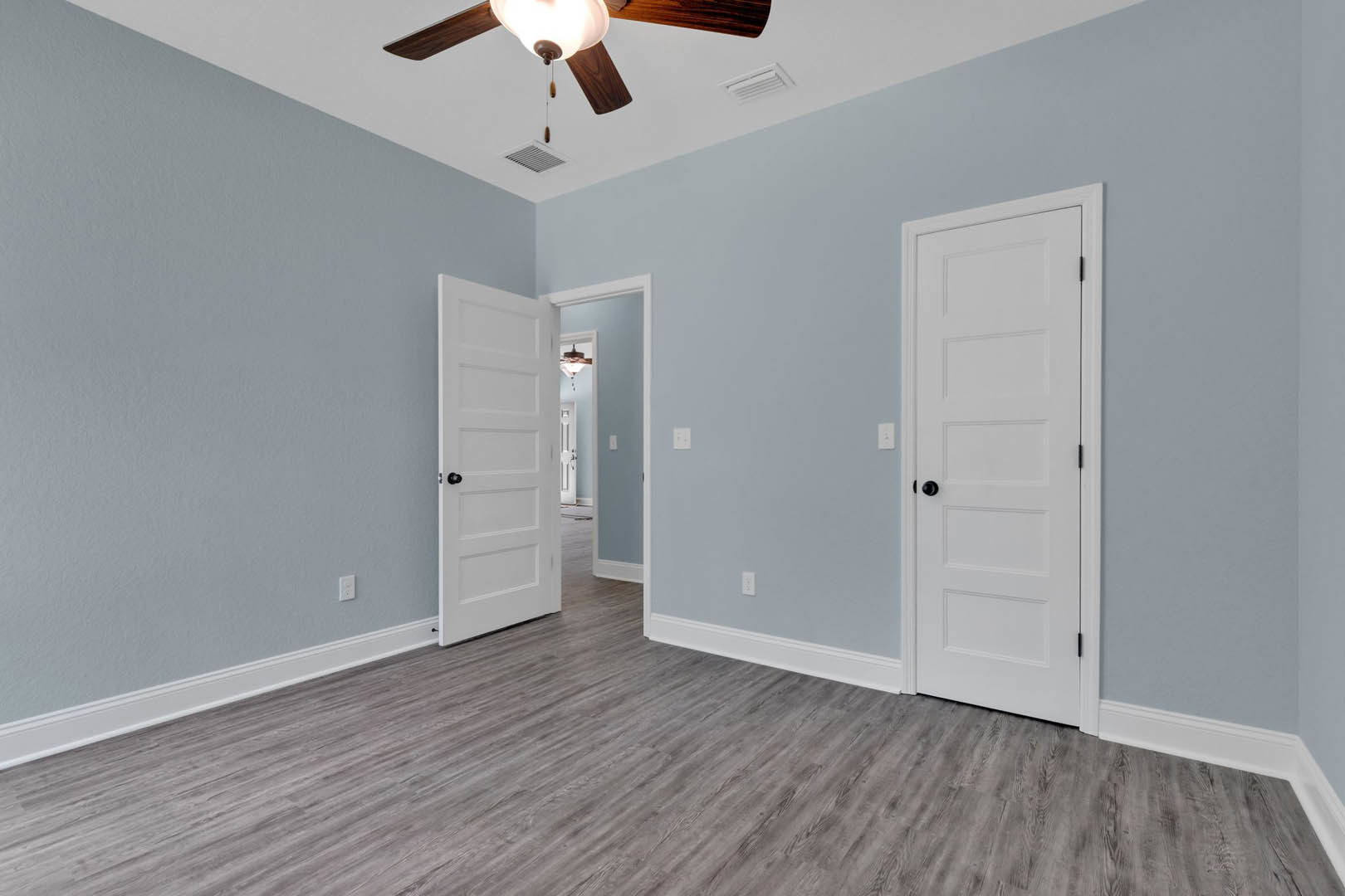Room with wood flooring and white trim, two white doors with black hardware, ceiling fan with light fixture on white plaster ceiling