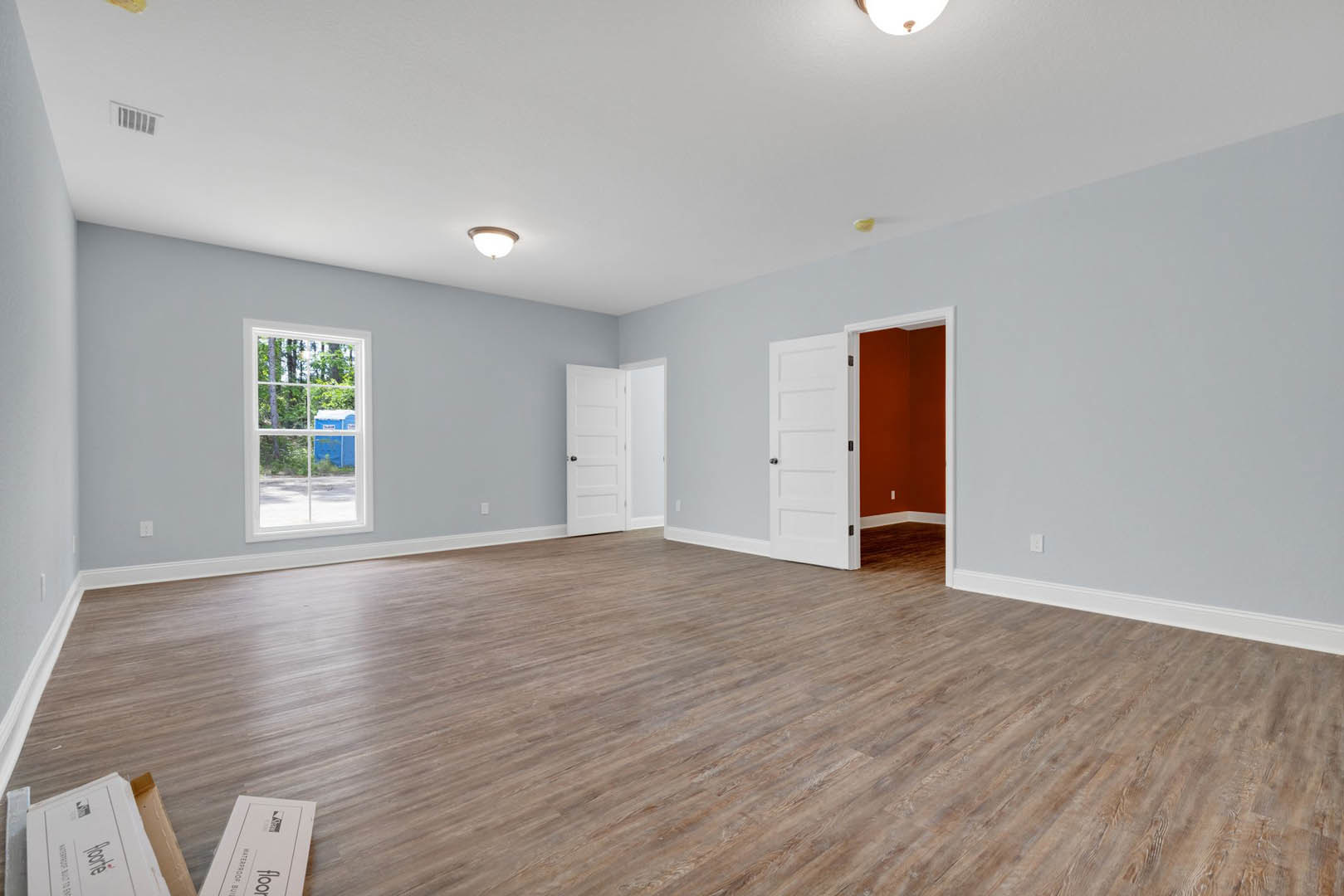 Wood floor room with white doors featuring black knobs, large window overlooking a blue container, ceiling-mounted light fixture, and white wall box with black text.