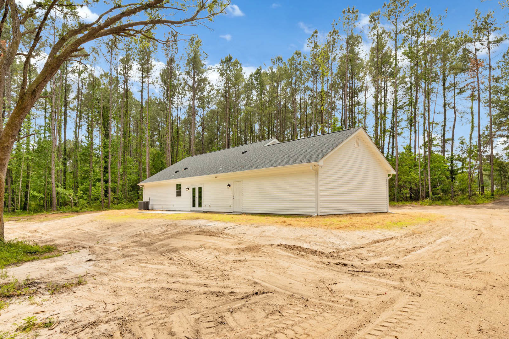 Modern house with black roof and white door, surrounded by dirt lot and mature trees under partly cloudy sky