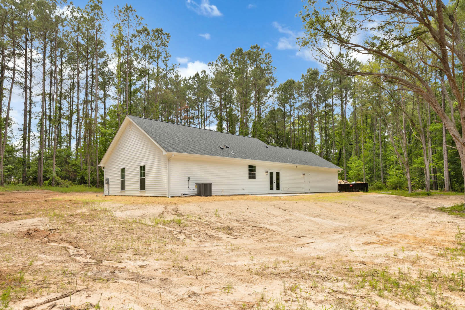 Modern cottage-style home surrounded by tall trees, blue sky overhead, grassy dirt lot in foreground, natural wood and stone exterior finishes.