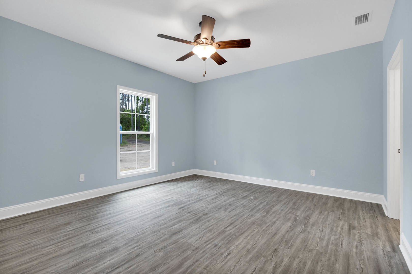 Ceiling fan with light fixture above wood laminate flooring, large window framed by white plaster walls, view of trees outside