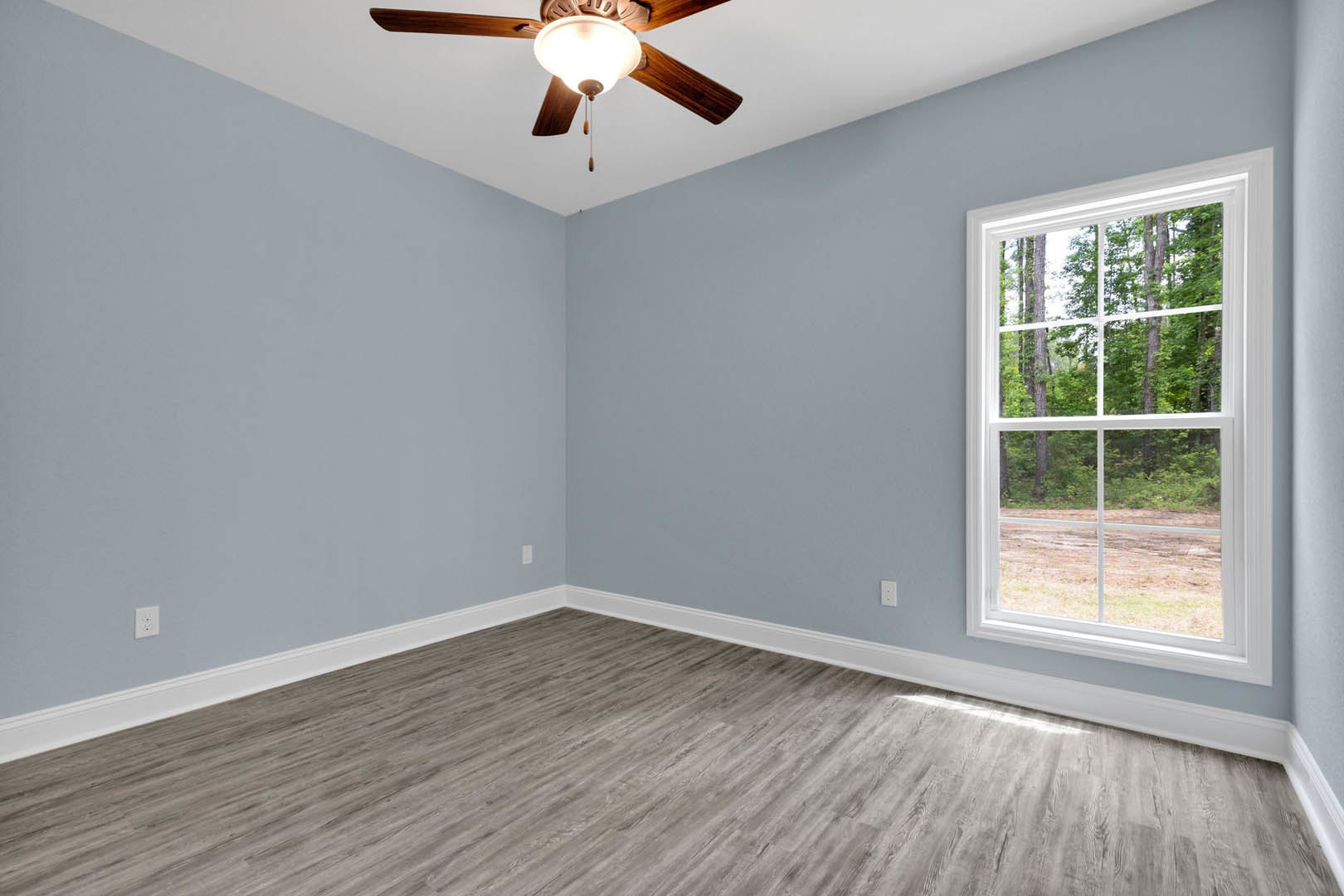 Wood flooring with white baseboards, ceiling fan with light fixture, large window overlooking trees, neutral plaster walls