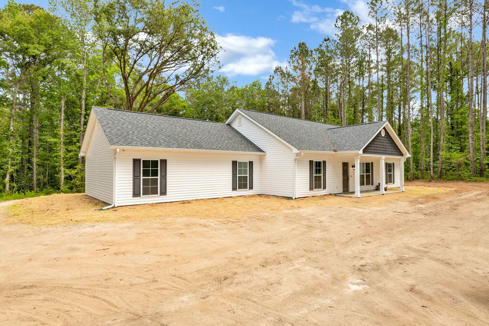 White house with brown shutters, white-framed windows, and a gabled roof, set beside a dirt road with trees and cloudy sky in the background