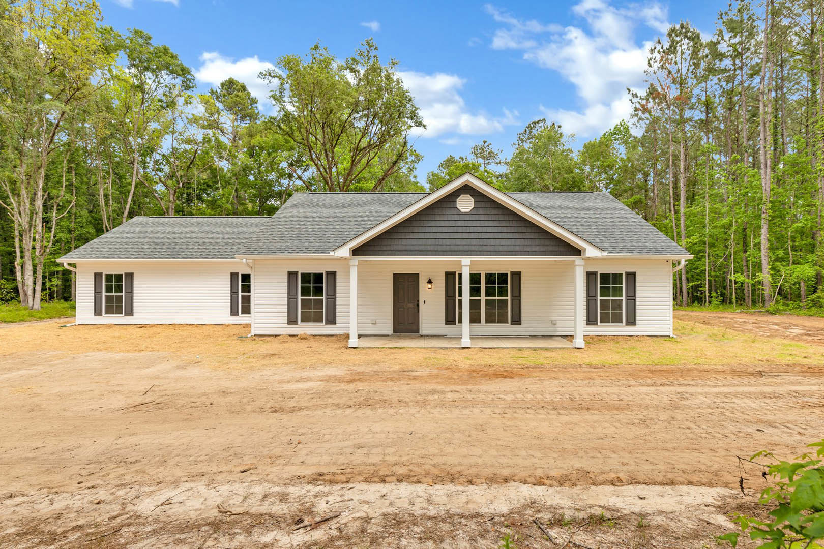 White siding house with front porch, illuminated entry light, white framed windows, dirt road leading up, surrounded by trees and cloudy sky in background