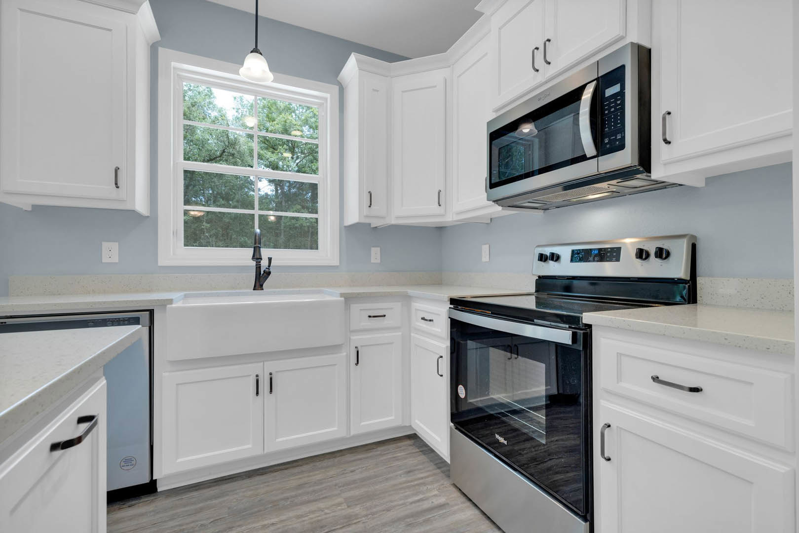 White kitchen cabinets, stainless steel sink, microwave above cabinetry, stove with glass oven door, window overlooking trees, white door with silver handle