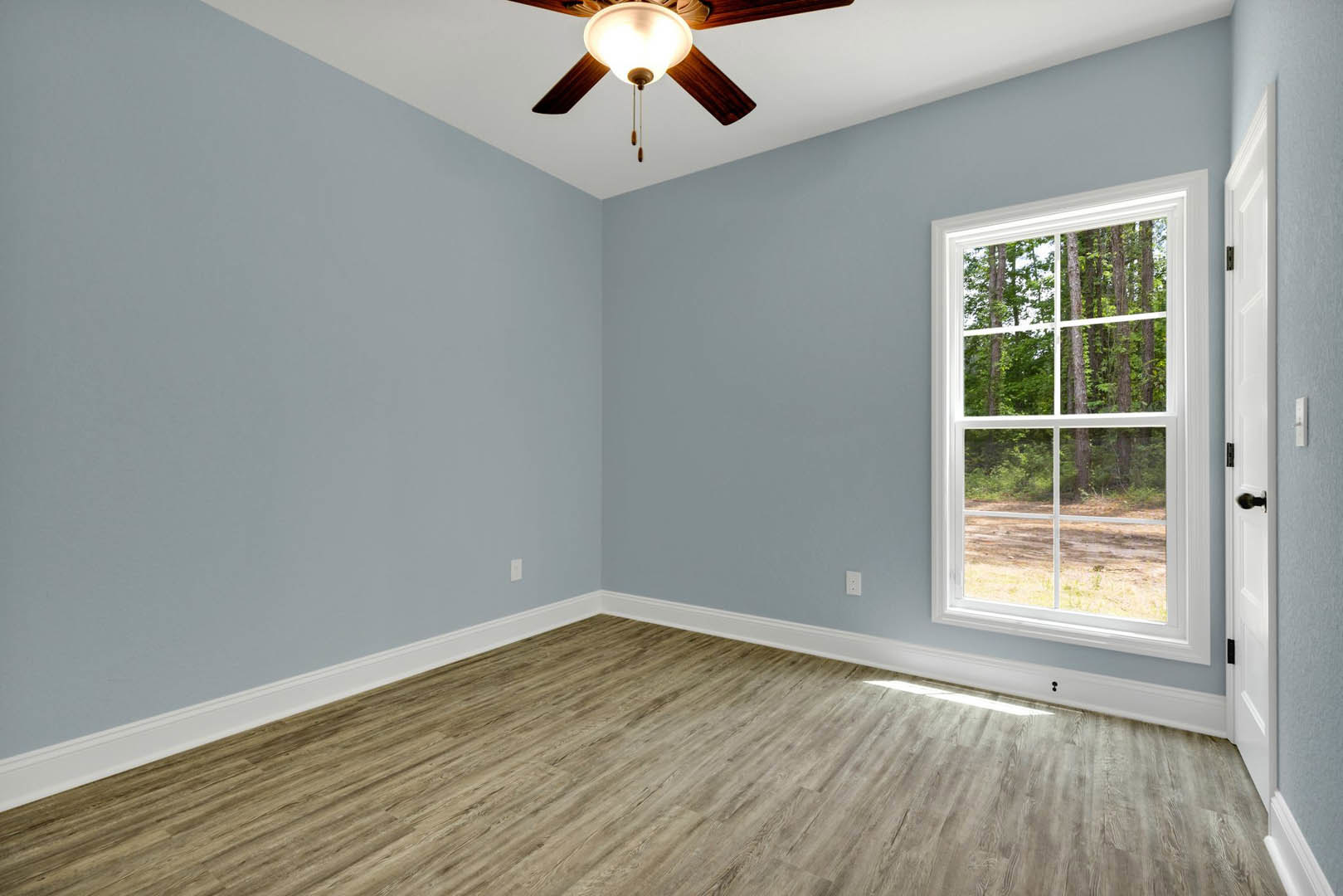 White-walled room with wood flooring, large window overlooking trees, white baseboards, and ceiling fan with light fixture