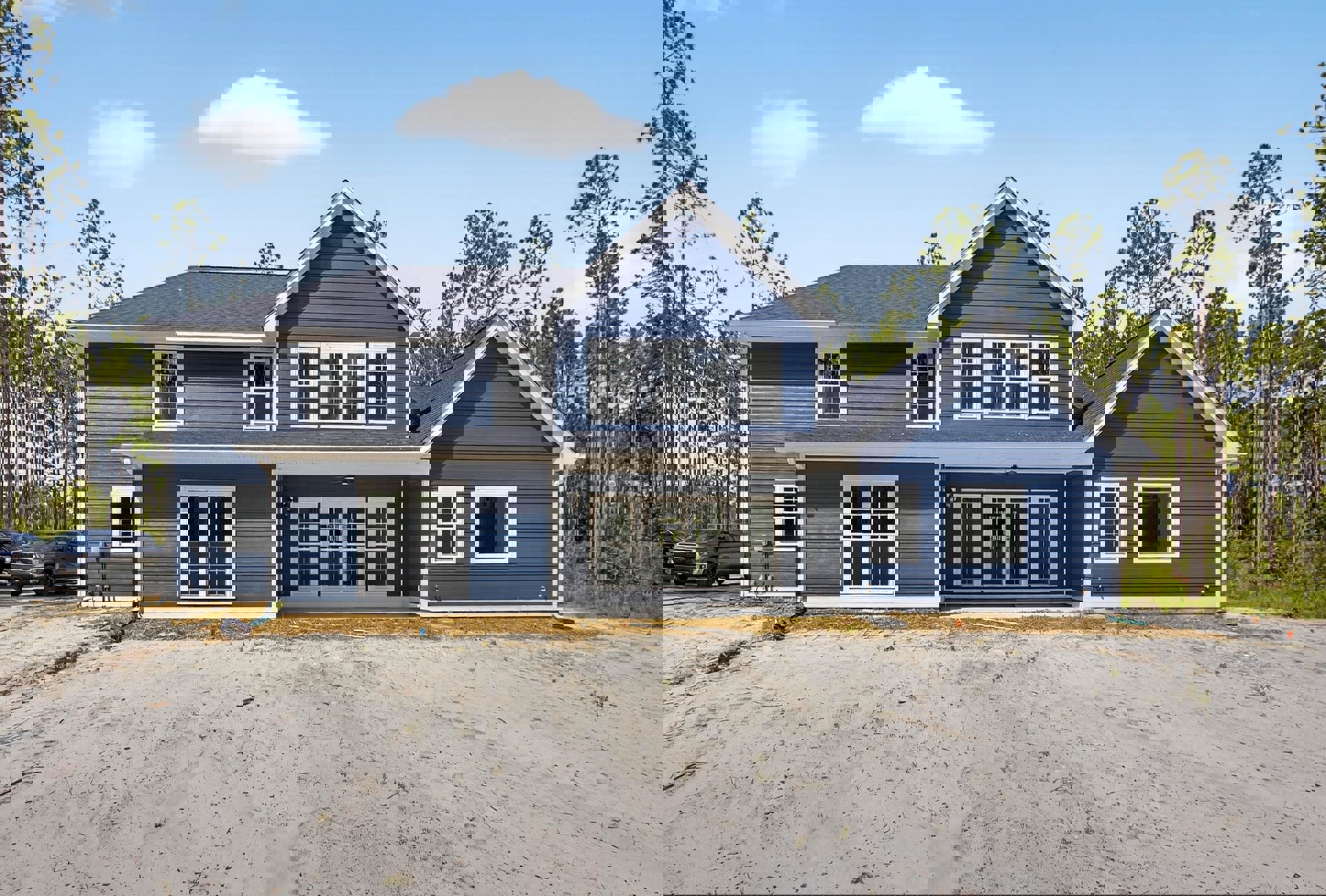 Blue house with white trim, gravel driveway, black truck parked nearby, large windows, front porch, surrounded by trees under a partly cloudy sky.