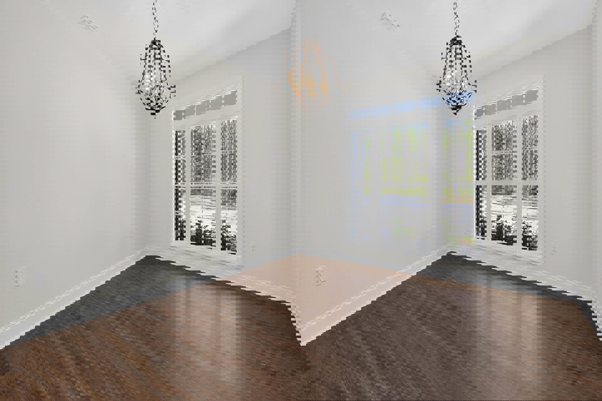 Chandelier with lit bulbs hangs above polished hardwood floor; large window reveals leafy trees outside; white electrical outlet and small white object rest on smooth white wall.