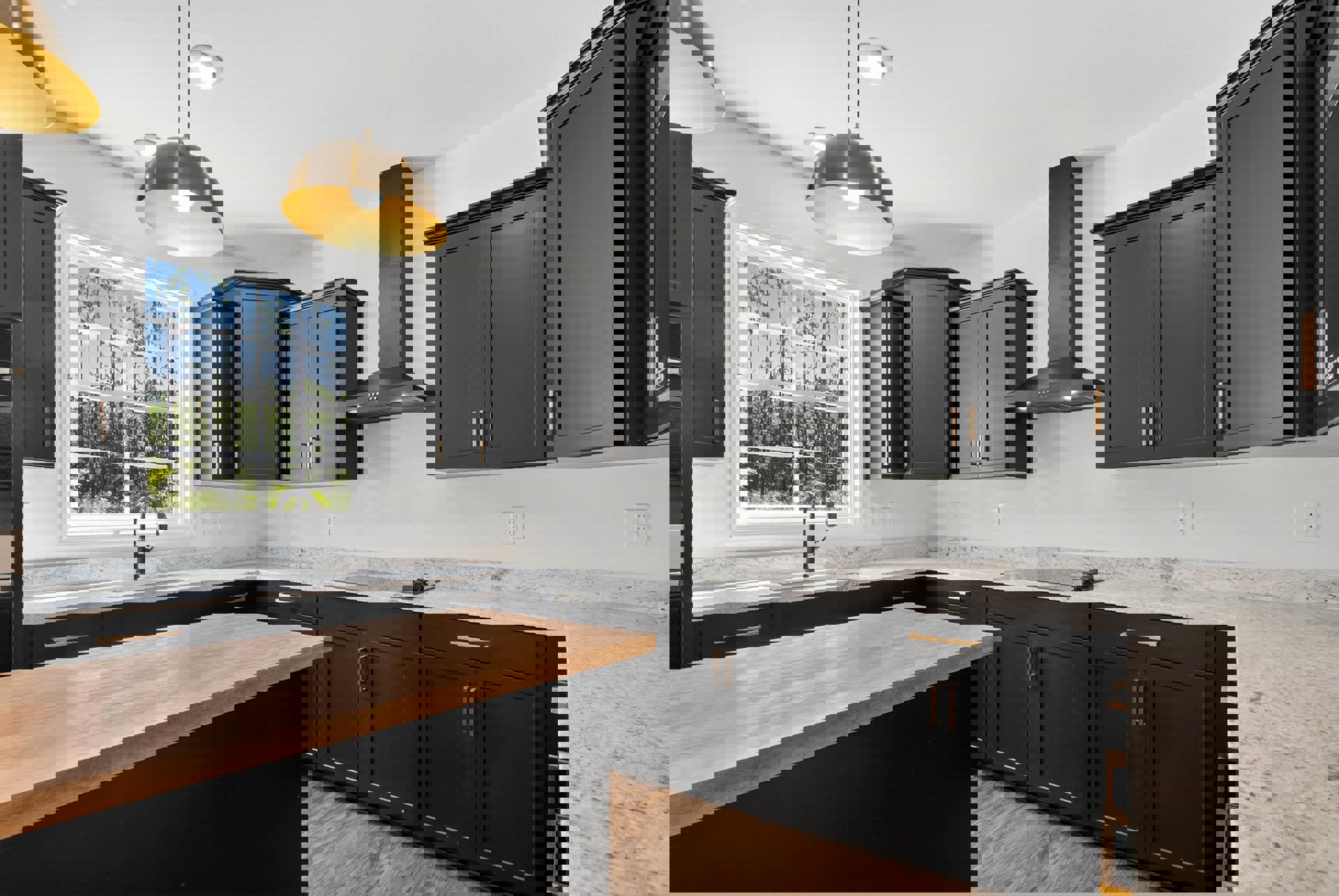 Black kitchen cabinets with gold handles, wood countertop, stainless steel faucet, pendant light fixture, window overlooking trees, close-up of dining table.