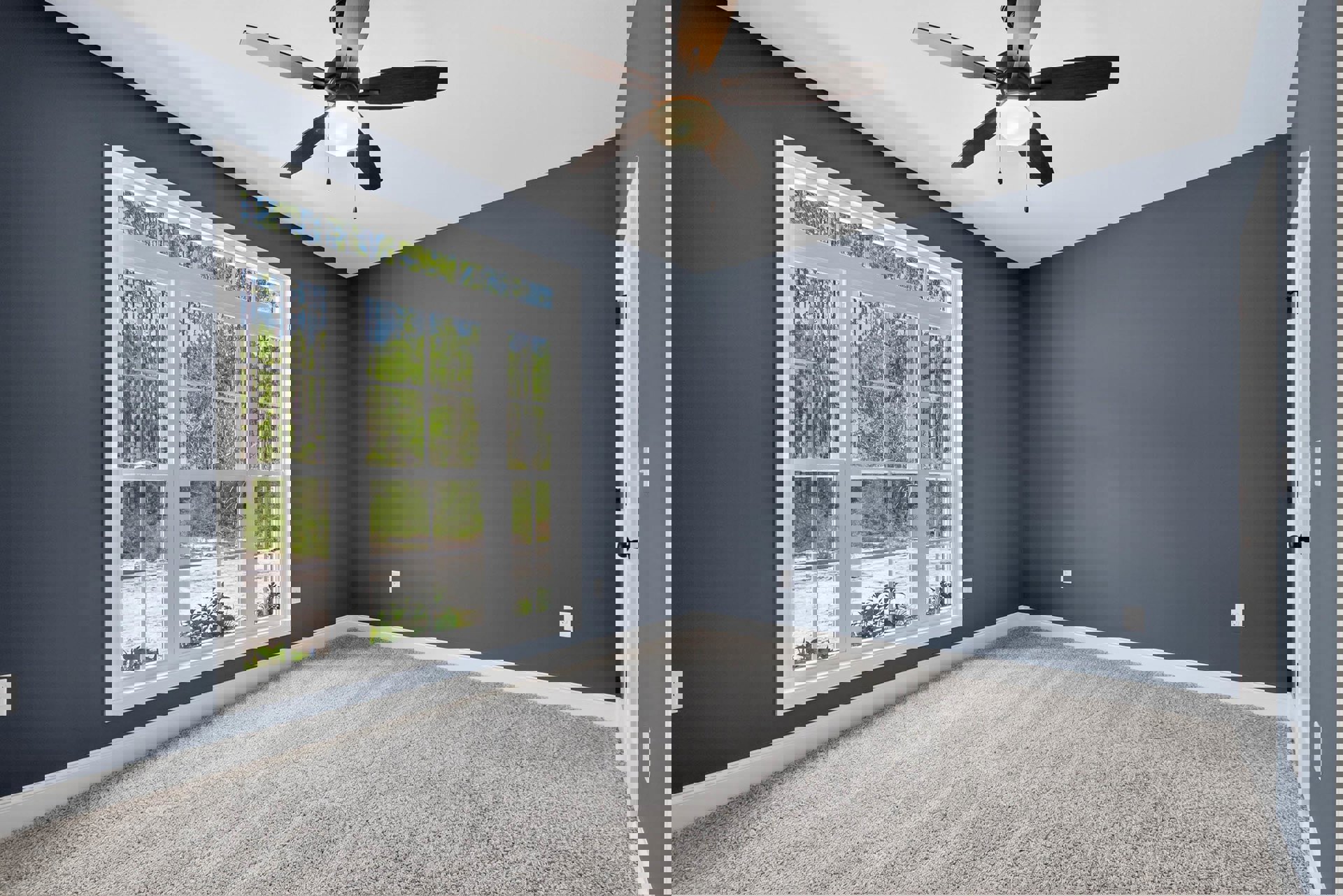 Neutral-toned carpeted room with white plaster walls, ceiling fan with integrated light, large window framing leafy trees, and a potted plant near the window.