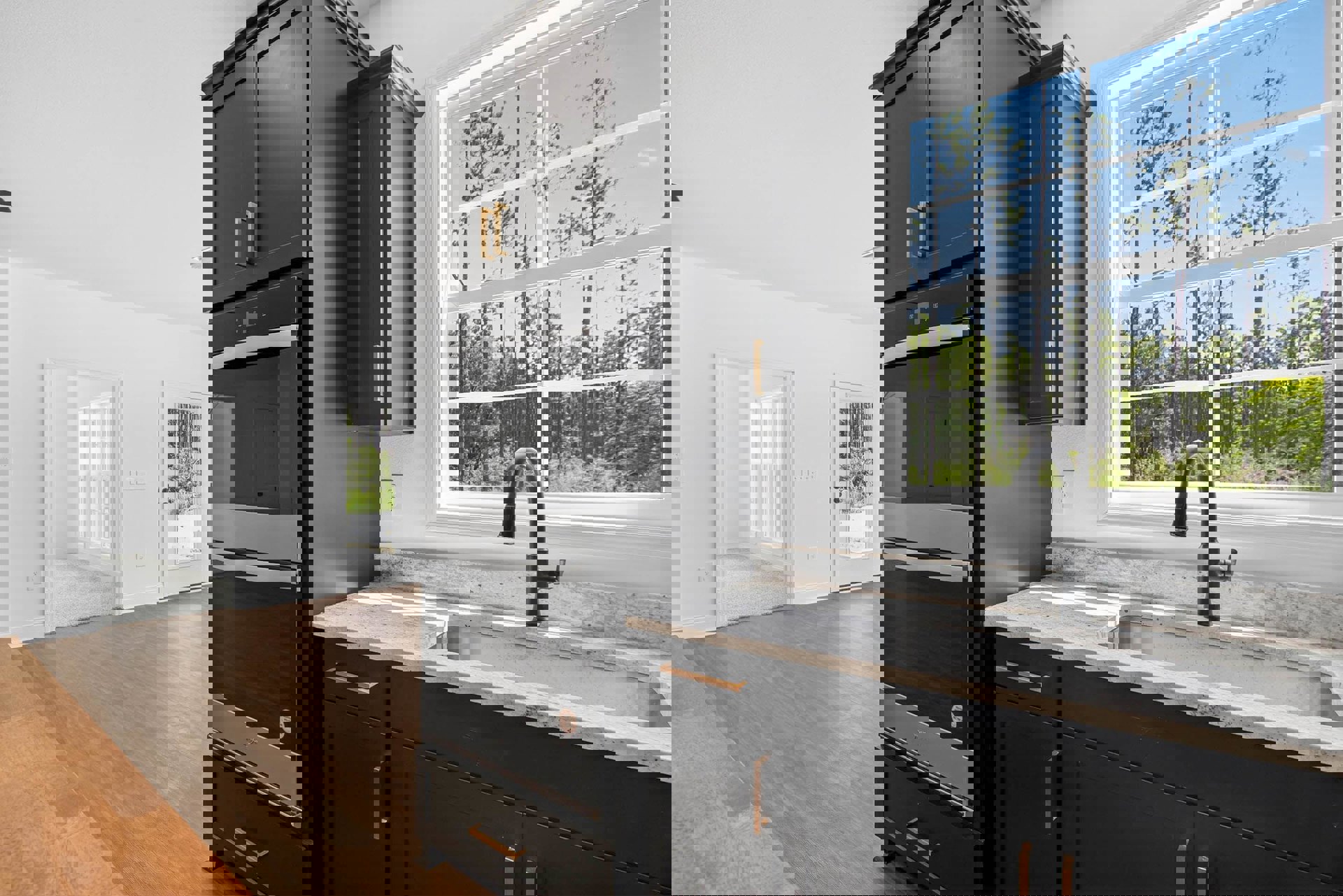 Black kitchen cabinets with marble countertops, stainless steel sink, black oven, black and silver refrigerator, hardwood flooring, white walls, window and door with views of trees