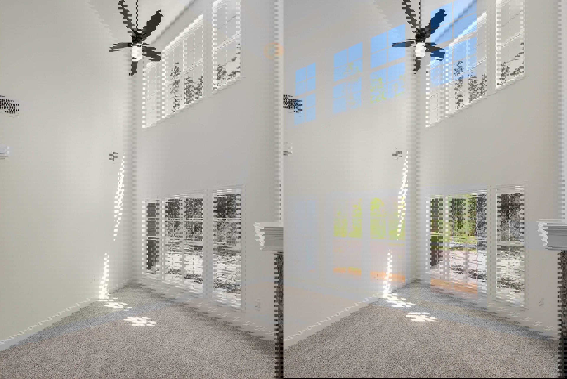 Neutral-toned carpeted room with white walls, ceiling fan with light fixture, and large windows overlooking green trees