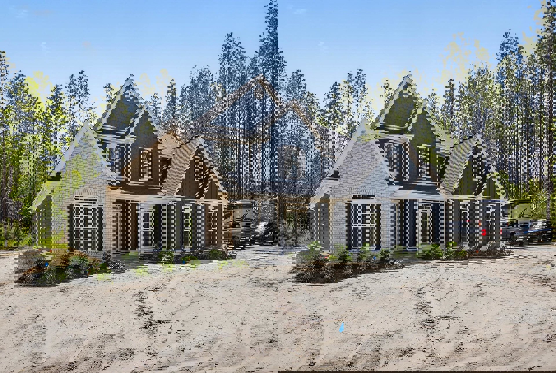 Two-story custom home with white windows, blue sign in sandy yard, police car with blue stripe, truck and camper trailer parked nearby, trees in background.