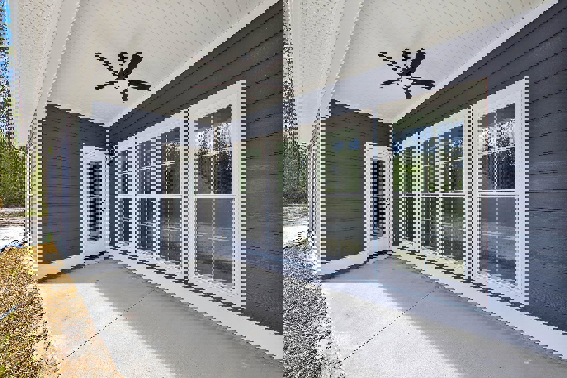 Covered porch with ceiling fan, row of windows, white door with glass insert, concrete floor, and view of trees and fence