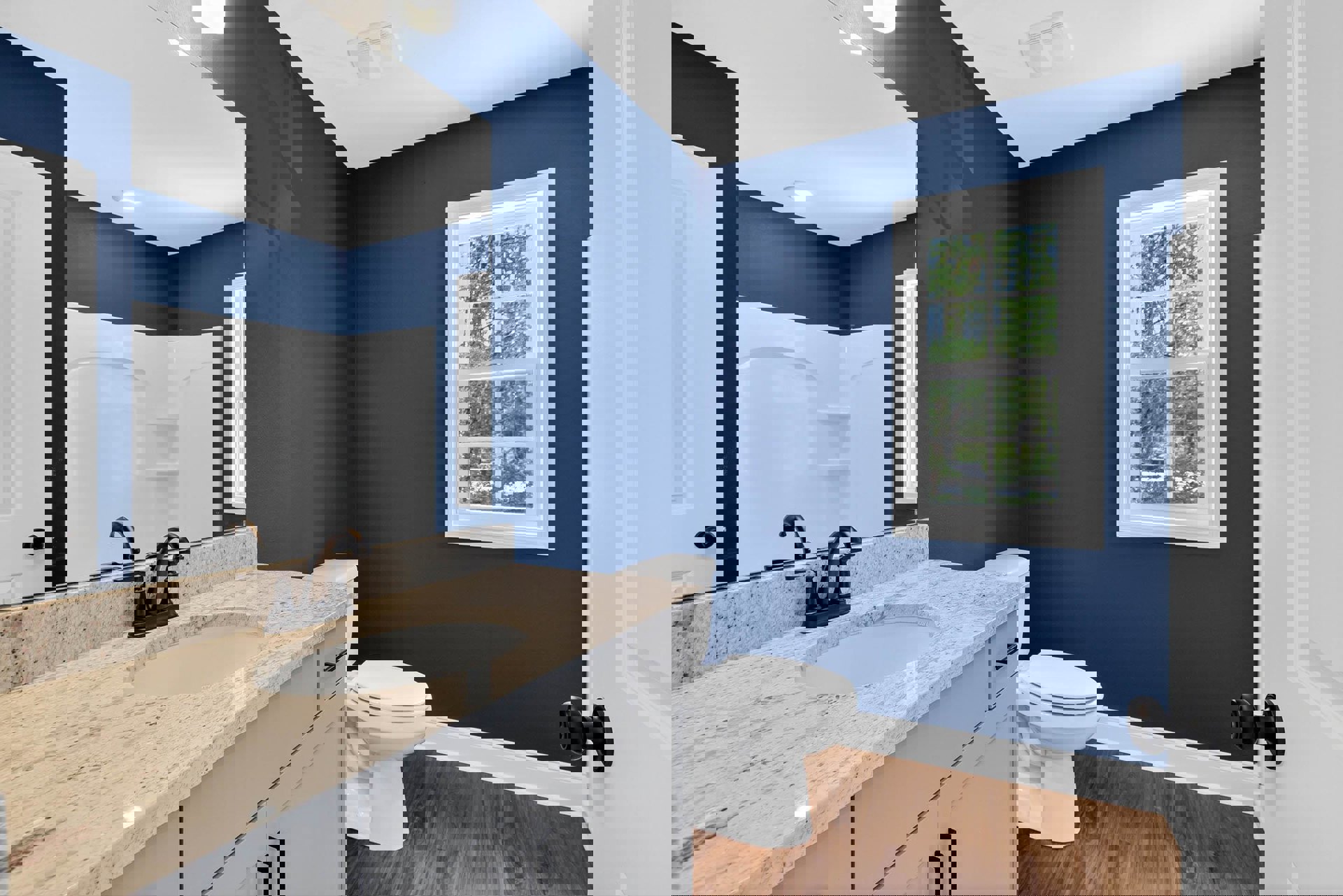 Bathroom with marble countertop, rectangular mirror above sink, white cabinetry, black faucet, white shower with black fixtures, closed toilet, window showing trees outside