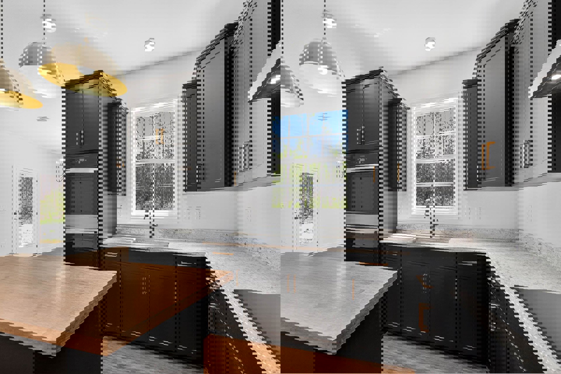 Black kitchen cabinets with matte finish, wooden dining table with black base, large window showing trees and blue sky, pendant light fixture, door opening to forest view