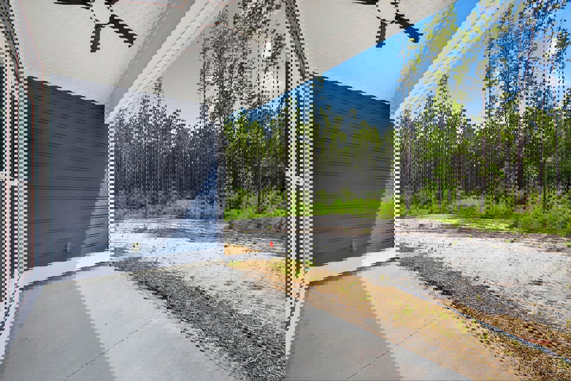 Covered porch with ceiling fan, blue siding and white trim, concrete driveway bordered by grass, dirt path leading to house, mature trees in background